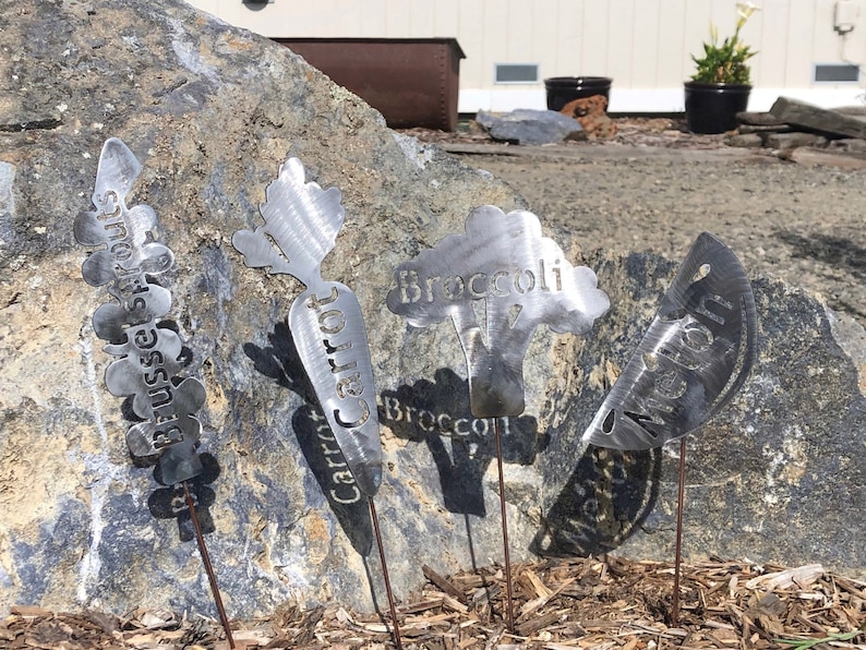 May include: Five metal garden markers with different vegetable names. The markers are shaped like the vegetables they represent. The names are "Brussels Sprouts", "Carrot", "Broccoli", "Melon", and "Watermelon".