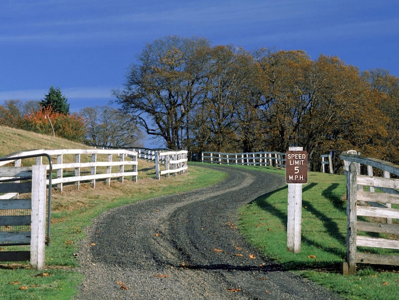 May include: A white wooden fence with a gate leads to a gravel driveway. A brown sign with white text reads "Speed Limit 5 M.P.H."