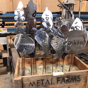 May include: A wooden crate labeled "METAL FARMS" holds a collection of metal garden markers. The markers are shaped like various vegetables and fruits, including a tomato, pepper, pumpkin, and chili pepper. Each marker has a metal rod attached to the bottom for inserting into the ground.