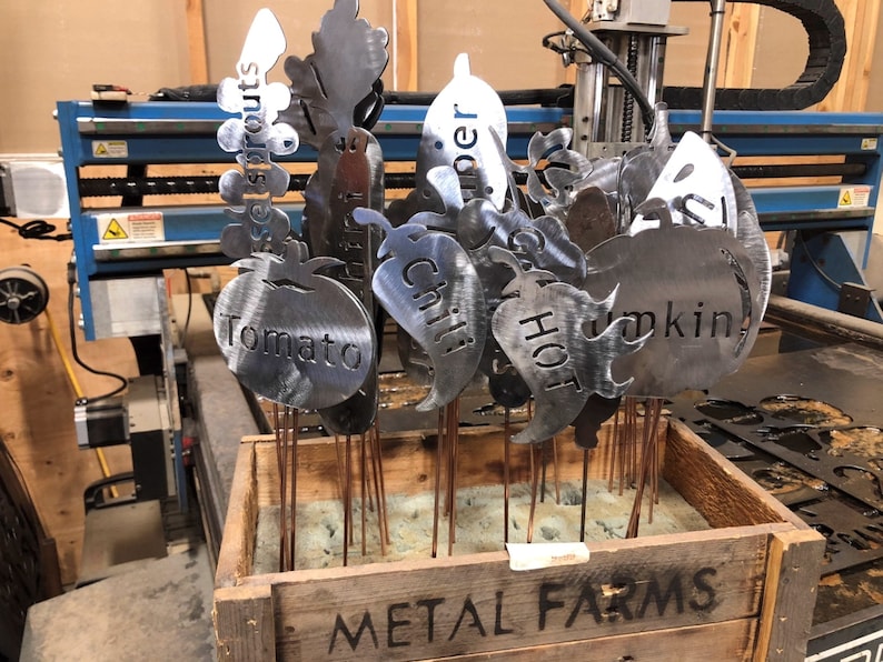 May include: A wooden crate labeled "METAL FARMS" holds a collection of metal garden markers. The markers are shaped like various vegetables and fruits, including a tomato, pepper, pumpkin, and chili pepper. Each marker has a metal rod attached to the bottom for inserting into the ground.