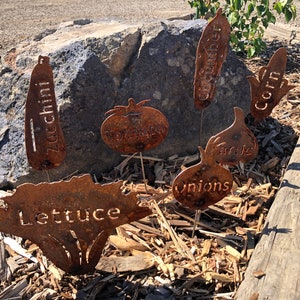 May include: Rustic metal garden markers with the names of vegetables: Zucchini, Tomato, Cucumber, Corn, Garlic, Onions, and Lettuce.