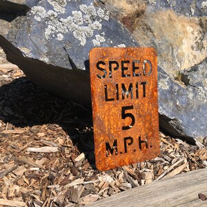 May include: A rusty metal sign with the text "SPEED LIMIT 5 M.P.H." The sign is sitting on a bed of wood chips.