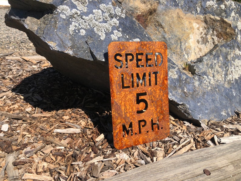 May include: A rusty metal sign with the text "SPEED LIMIT 5 M.P.H." The sign is sitting on a bed of wood chips.