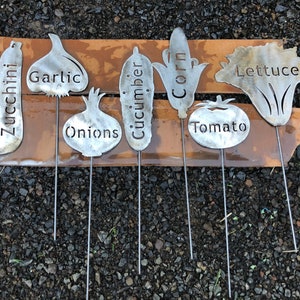 May include: Set of eight metal garden markers shaped like vegetables with the names of the vegetables written on them: zucchini, garlic, onions, cucumber, corn, tomato, and lettuce.