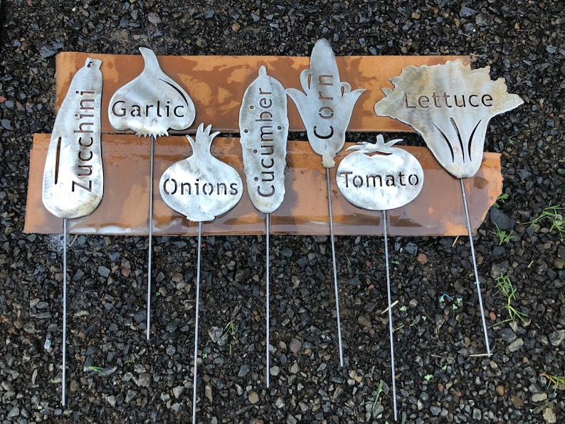 May include: Set of eight metal garden markers shaped like vegetables with the names of the vegetables written on them: zucchini, garlic, onions, cucumber, corn, tomato, and lettuce.