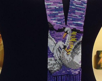 May include: A purple, blue, and white beaded necklace with a beaded image of two swans and a yellow sun. The necklace has a fringe of purple beads.