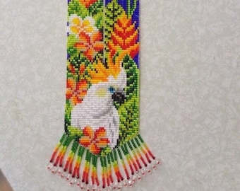 May include: A beaded necklace featuring a white cockatoo, orange, yellow, and green flowers, and a blue and green background. The necklace has a fringe of beads in various colours.