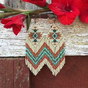 Op de afbeelding: Een paar lange, geperde oorbellen met een geometrisch patroon in beige, bruin, turquoise en oranje tinten. De oorbellen hebben een franje-achtig ontwerp aan de onderkant.