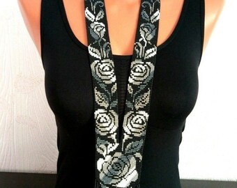 May include: A beaded necklace featuring a floral design. The necklace is primarily black with white, gray, and blue rose patterns. The necklace has a long, fringed bottom and is displayed on a mannequin.