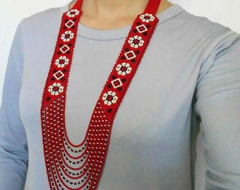 May include: A long, multi-strand beaded necklace with a red and white geometric pattern. The necklace is made of small, round beads and has a tassel at the bottom.