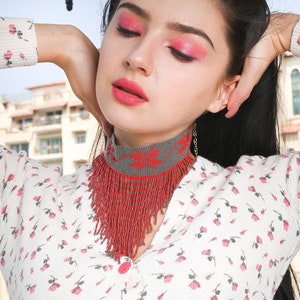 Peut inclure: Une femme portant une blouse blanche &agrave; motif floral et un collier ras du cou en perles rouges et grises. Le collier a un design en frange et est superpos&eacute; &agrave; une cha&icirc;ne argent&eacute;e.
