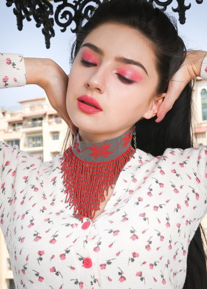 Peut inclure: Une femme portant une blouse blanche &agrave; motif floral et un collier ras du cou en perles rouges et grises. Le collier a un design en frange et est superpos&eacute; &agrave; une cha&icirc;ne argent&eacute;e.