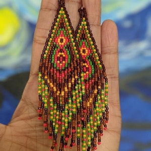 May include: A pair of long, beaded earrings with a brown, green, yellow, and red colour scheme. The earrings are made of small, colourful beads that are strung together in a geometric pattern. The earrings are hanging from a pair of gold-coloured hooks.