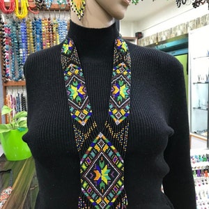 Könnte beinhalten: Eine lange, farbenfrohe Perlenkette mit geometrischem Muster und Fransen. Die Kette besteht aus schwarzen, orangefarbenen, gelben, grünen, blauen und weißen Perlen. Die Kette wird an einem Mannequin mit einem schwarzen Pullover präsentiert.