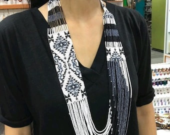 May include: A long, white and black beaded necklace with a geometric pattern. The necklace is layered and has a fringe at the bottom. The necklace is paired with matching earrings.