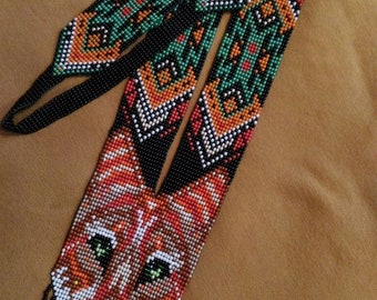 May include: A beaded necklace featuring a fox face design. The fox is made with orange, white and black beads. The necklace has a geometric pattern with green, orange and black beads.