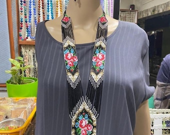May include: A long black beaded necklace with a floral design and fringe. The necklace is made of small black beads with white and gold accents. The floral design is in shades of pink, red, blue, and green. The necklace is displayed on a mannequin wearing a gray shirt.