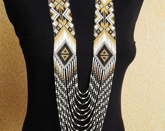 May include: A long, multi-strand beaded necklace with a geometric pattern in black, white, and gold. The necklace is made of small, round beads and has a cascading effect.
