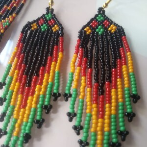 May include: A pair of long, beaded earrings with a black, red, yellow, and green colour scheme. The earrings are made of small, round beads and have a tassel-like design.