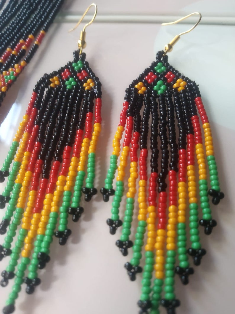 May include: A pair of long, beaded earrings with a black, red, yellow, and green colour scheme. The earrings are made of small, round beads and have a tassel-like design.