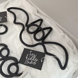May include: Two black knitted bat shapes on a white background with black polka dots. The bats are made of thick rope and are hanging from the ceiling. A black tag with white text reads "hey kiddo studio".