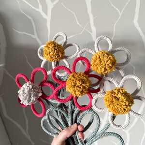 Op de afbeelding: Een boeket van vijf gehaakte bloemen met pompoenen in tinten wit, grijs en geel. De bloemen zijn verbonden door een grijze gehaakte steel.