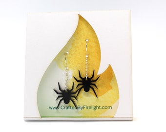 May include: Black spider earrings on a white card with a yellow flame design. The earrings are on silver chains and have a black spider charm.