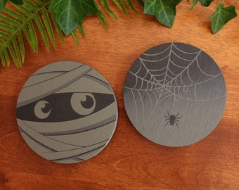 Puede incluir: Dos posavasos redondos de pizarra con diseños de Halloween. Un posavasos presenta una momia gris y negra con ojos. El otro posavasos presenta una telaraña negra con una araña negra en el centro.