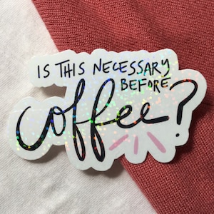 May include: A holographic sticker with the text "IS THIS NECESSARY BEFORE coffee?" in black script. The sticker is white with a colorful, iridescent sheen and is placed on a white and red-orange fabric background.