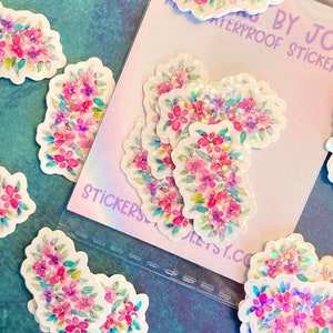 May include: A collection of floral stickers with a watercolor design. The stickers feature pink and purple flowers with green leaves and iridescent accents. The text "STICKERS BY JODI WATERPROOF STICKERS" is visible on the packaging.