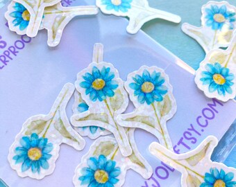 May include: A collection of daisy flower stickers with blue petals and yellow centers. The stickers have a watercolor effect and are on a white background. The stems and leaves are light green. The stickers are scattered on a light blue surface.