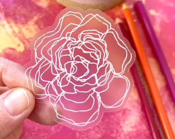 May include: A translucent sticker featuring a white outline of a peony flower. The sticker has a gradient of pink and orange hues. The image also includes colored pencils and a pink mug.