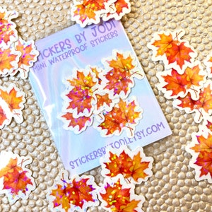 May include: Mini waterproof stickers featuring vibrant orange and yellow maple leaves with glitter accents. The stickers are packaged in a clear plastic sleeve with the text "Stickers by Jodi" and "Mini Waterproof Stickers".