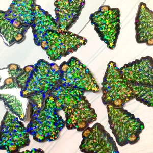 May include: A collection of Christmas tree-shaped stickers with a glittery, holographic effect. The trees are primarily green with accents of blue, yellow, and red, and have a dark brown border. Some trees have a small yellow star at the top.