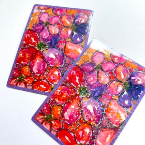 May include: Two rectangular playing cards with a floral design. The artwork features a watercolor style with red, orange, purple, and blue flowers. The edges of the cards are purple, and the background is white.