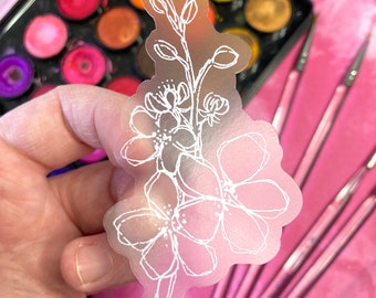 May include: A translucent sticker featuring a white floral design. The sticker showcases a delicate outline of flowers and leaves. The background includes a watercolor palette and pink paintbrushes, suggesting an art or craft theme.