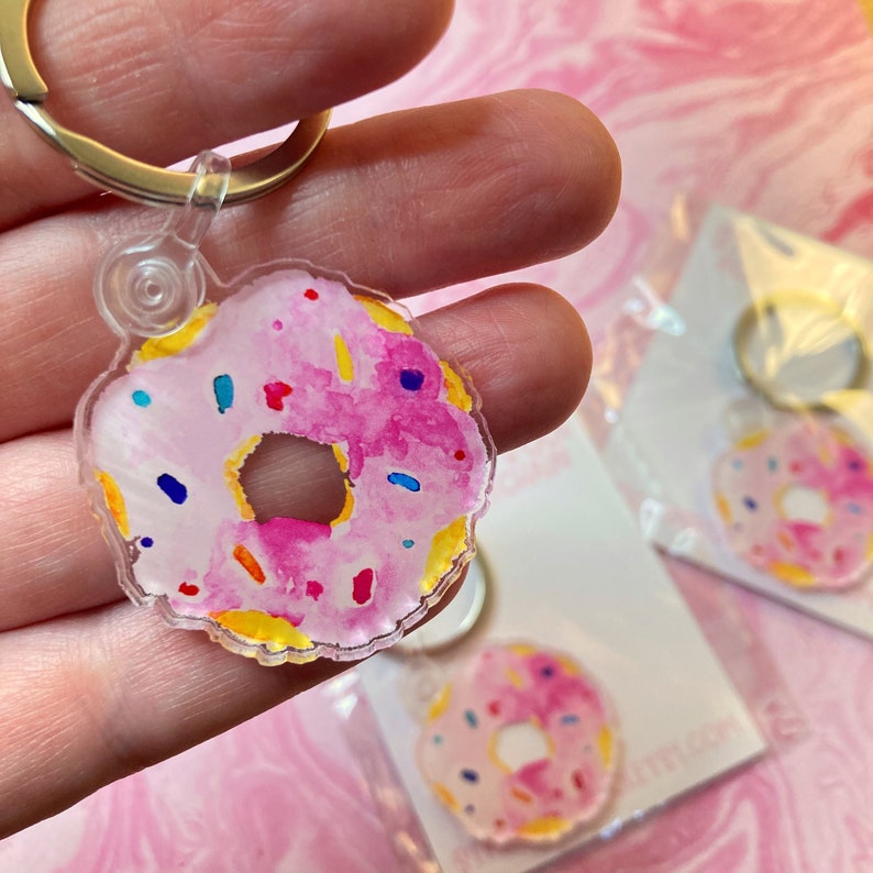 Puede incluir: Un llavero transparente en forma de rosquilla de acr&iacute;lico con un dise&ntilde;o de acuarela rosa y chispas coloridas. El llavero tiene un anillo dorado para sujetarlo a llaves o bolsos. El dise&ntilde;o de la rosquilla es visible en la parte delantera.