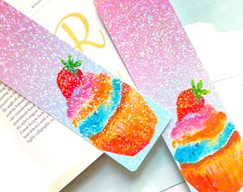 May include: Two glittery bookmarks with a cupcake design. The cupcakes have a red strawberry on top, with orange, blue, and pink frosting. The bookmarks have a pink glitter gradient.