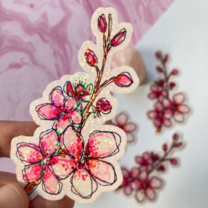 May include: A sticker featuring a watercolor illustration of a cherry blossom branch with pink flowers and buds. The sticker has a glittery, holographic finish and a black outline. Additional stickers are visible in the background.