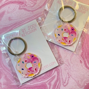 Puede incluir: Dos llaveros de acr&iacute;lico con forma de donut con un dise&ntilde;o de acuarela rosa y chispas coloridas. Cada llavero est&aacute; unido a un llavero plateado y empaquetado en una tarjeta blanca con el texto "STICKERS BY JODI KEYCHAIN" y una direcci&oacute;n web.