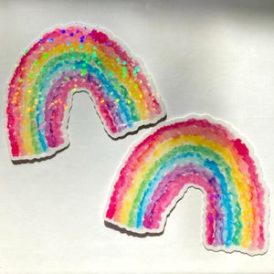 May include: Two rainbow-shaped resin coasters. Each coaster features a watercolor design with bands of red, orange, yellow, green, blue, and purple. One coaster has iridescent glitter accents. The coasters are on a white background.