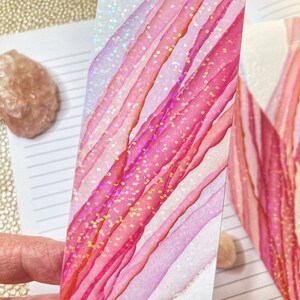 May include: A handmade bookmark with a vibrant abstract design. The bookmark features diagonal stripes in shades of pink and white, accented with iridescent glitter. The design is reminiscent of watercolor art, perfect for book lovers.