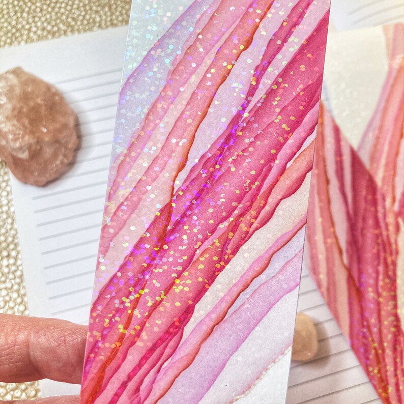 May include: A handmade bookmark with a vibrant abstract design. The bookmark features diagonal stripes in shades of pink and white, accented with iridescent glitter. The design is reminiscent of watercolor art, perfect for book lovers.