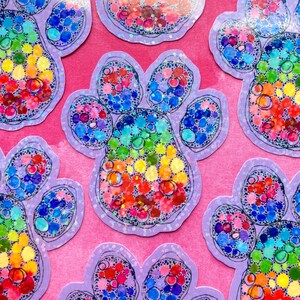 May include: A collection of colorful paw print stickers. Each sticker features a rainbow of colors within the paw print shape, outlined in black and set against a lavender background. The stickers are scattered across a pink surface.