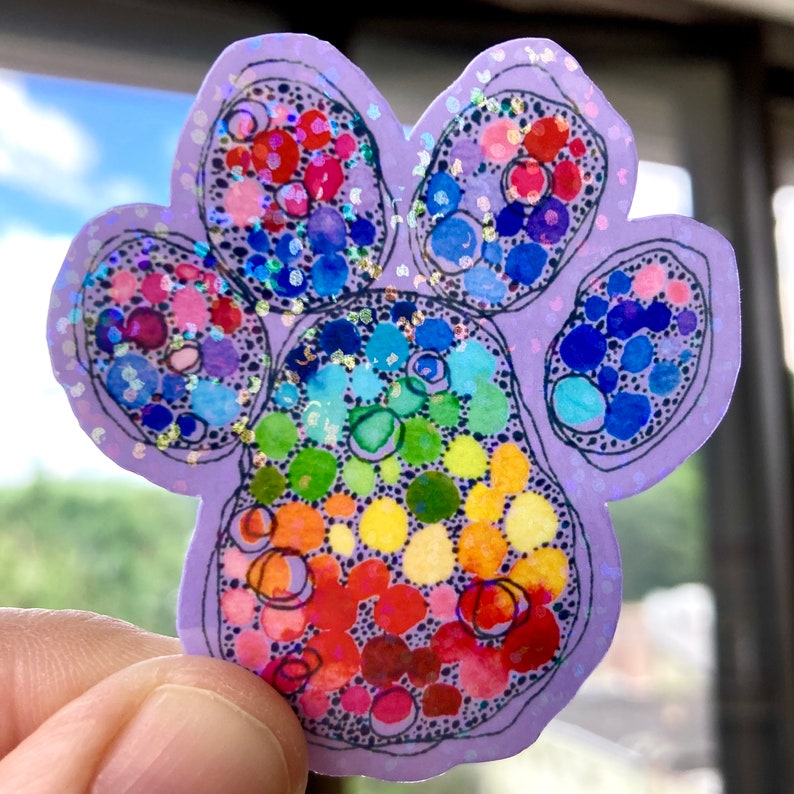 May include: A paw-shaped sticker with a rainbow-colored design. The sticker features a light purple outline and is filled with colorful circles and dots. The design includes red, orange, yellow, green, blue, and purple hues. The sticker has a holographic, glittery finish.
