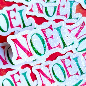 May include: A collection of glittery stickers with the word "NOEL" in a festive font. The letters are in shades of green and pink, with a shimmering, iridescent effect. The stickers are on a red background.