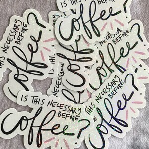 May include: A collection of white stickers with the text "Is this necessary before coffee?" in black script. The word "coffee" is in a large, stylized font. The stickers have a holographic, glittery effect with pink accents.