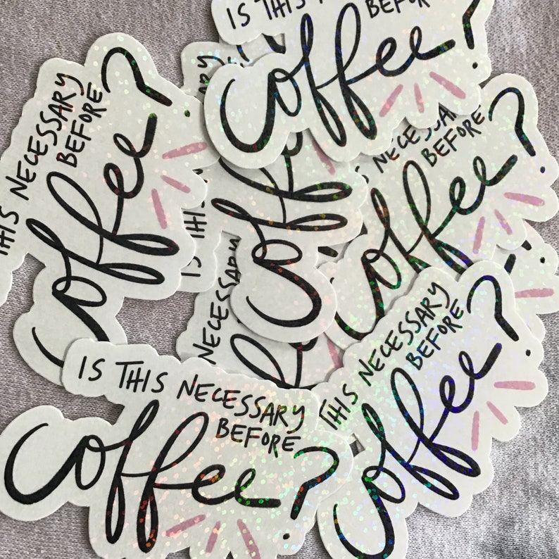 May include: A collection of white stickers with the text "Is this necessary before coffee?" in black script. The word "coffee" is in a large, stylized font. The stickers have a holographic, glittery effect with pink accents.