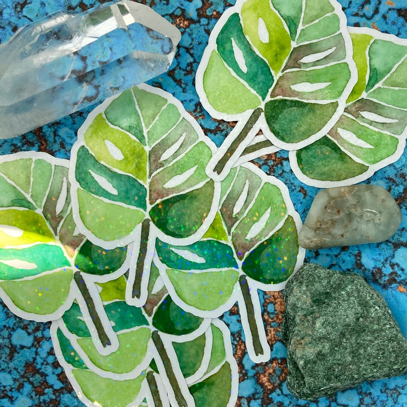 K&ouml;nnte beinhalten: Eine Sammlung von Aquarell-Blatt-Aufklebern in Gr&uuml;nt&ouml;nen mit wei&szlig;em Rand, auf einer strukturierten blauen Oberfl&auml;che. Klarer Quarz und verschiedene Steine erg&auml;nzen die Aufkleber und schaffen ein nat&uuml;rliches, botanisches Thema.