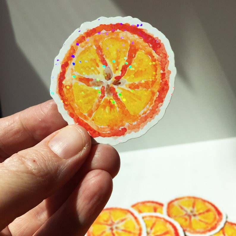 Puede incluir: Una pegatina de acuarela de una rodaja de naranja con un acabado brillante e iridiscente. La pegatina presenta un centro amarillo brillante, una c&aacute;scara rojo-naranja y un borde blanco. La pegatina est&aacute; sujeta por una mano.